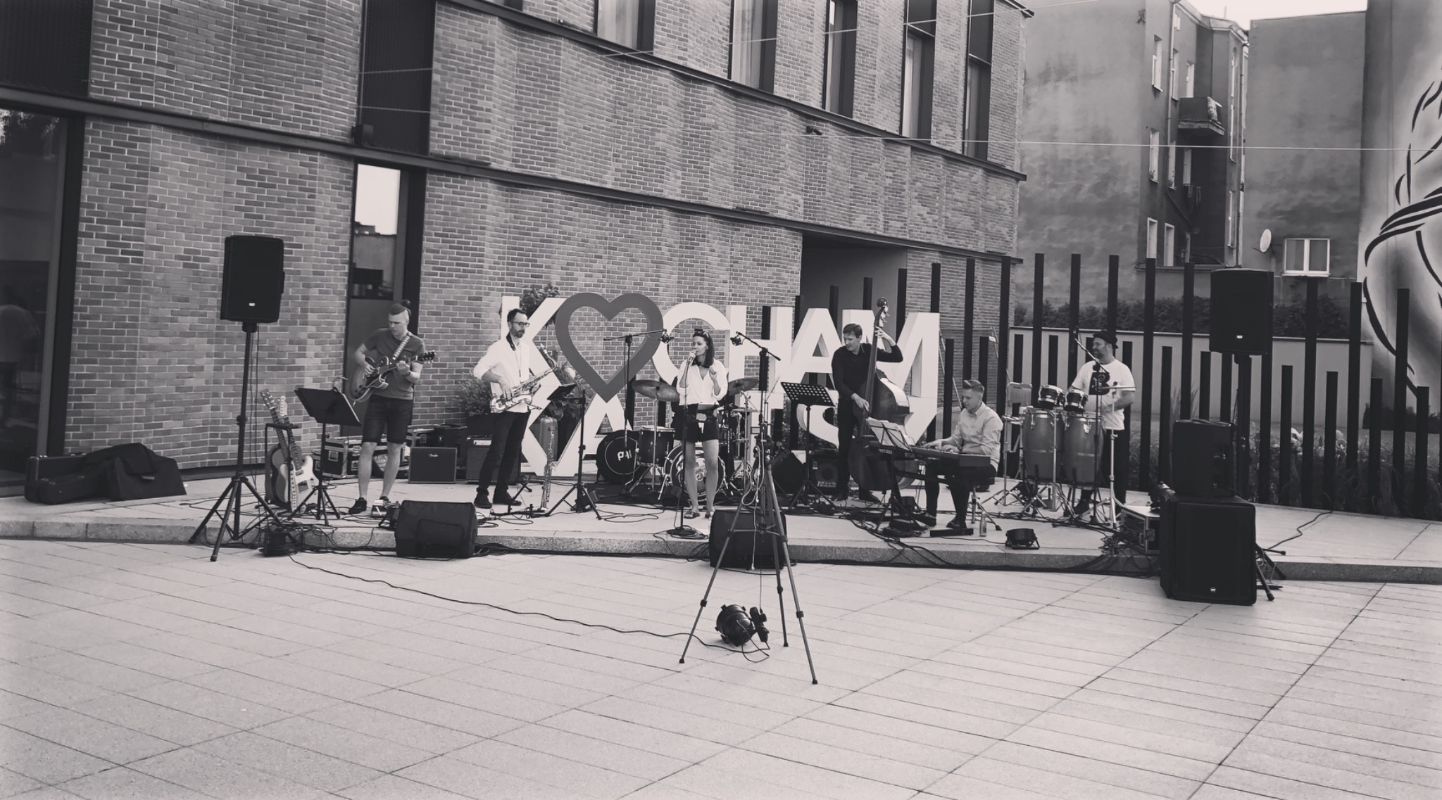 Zespół muzyczny grający na zewnątrz, z saksofonistą, gitarzystą, wokalistką, kontrabasistą, klawiszowcem i perkusistą, na tle ceglanego budynku i dekoracji z napisem 'KOCHAM'