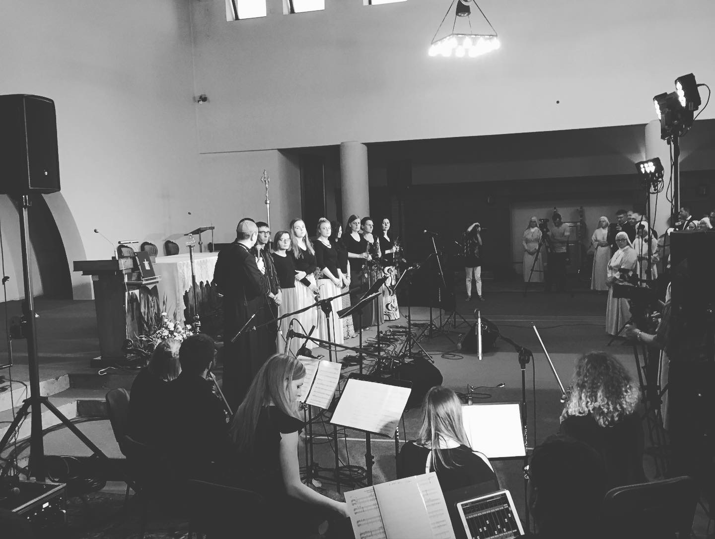 Czarno-białe zdjęcie chóru podczas występu w kościele, widoczne mikrofony, statywy i reflektory, w tle grupa zakonnic.