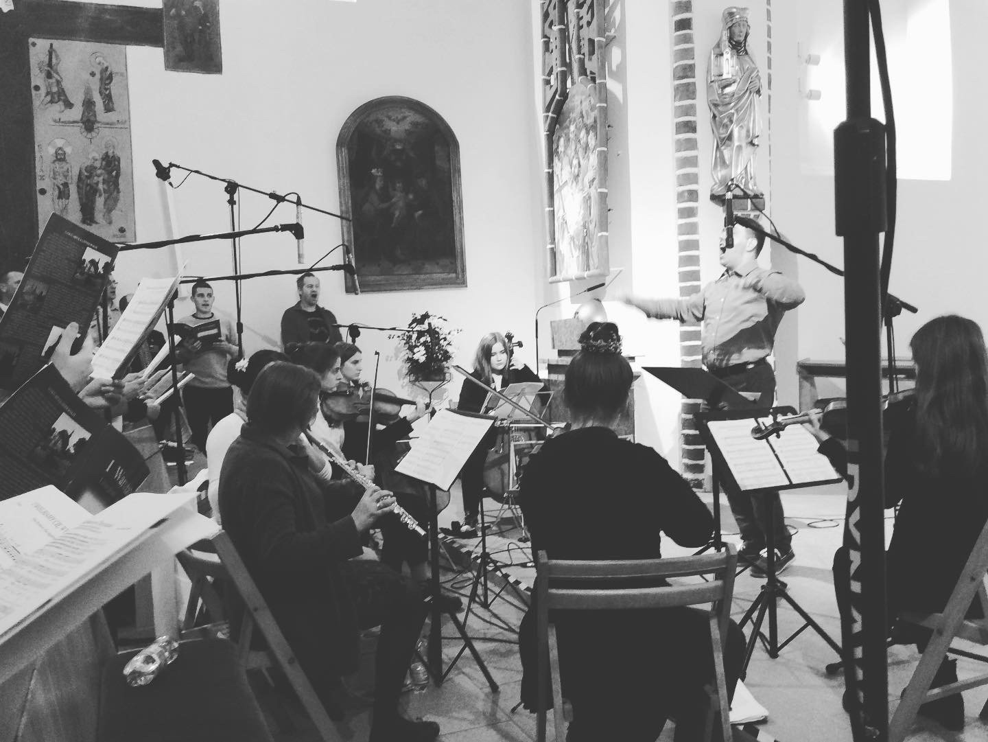 Czarno-białe zdjęcie chóru i orkiestry podczas występu w kościele, z mikrofonami skierowanymi w stronę śpiewaków i instrumentów.
