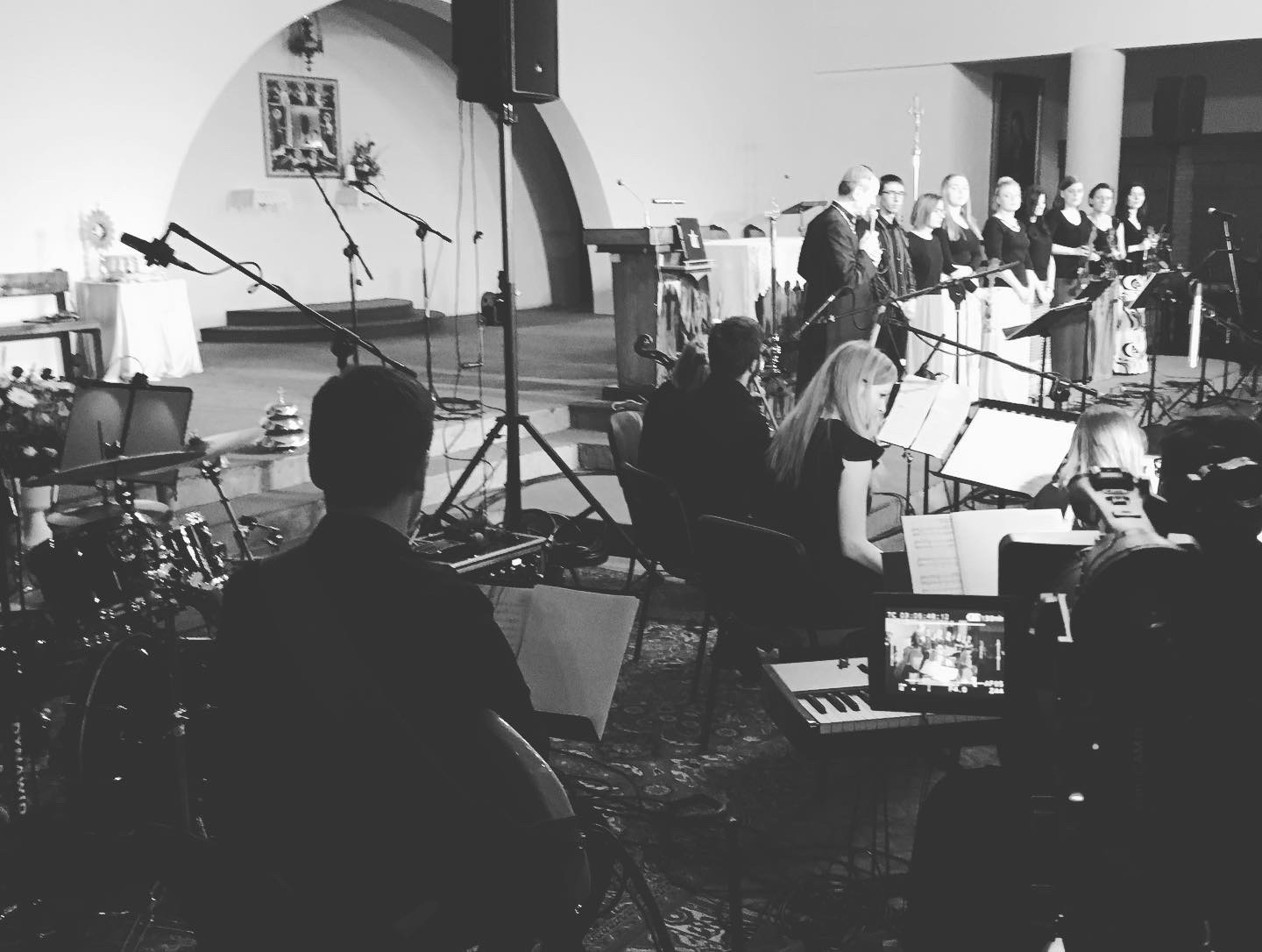 Czarno-białe zdjęcie zespołu muzycznego i chóru podczas występu w kościele, widoczne mikrofony, instrumenty i pulpit z nutami.