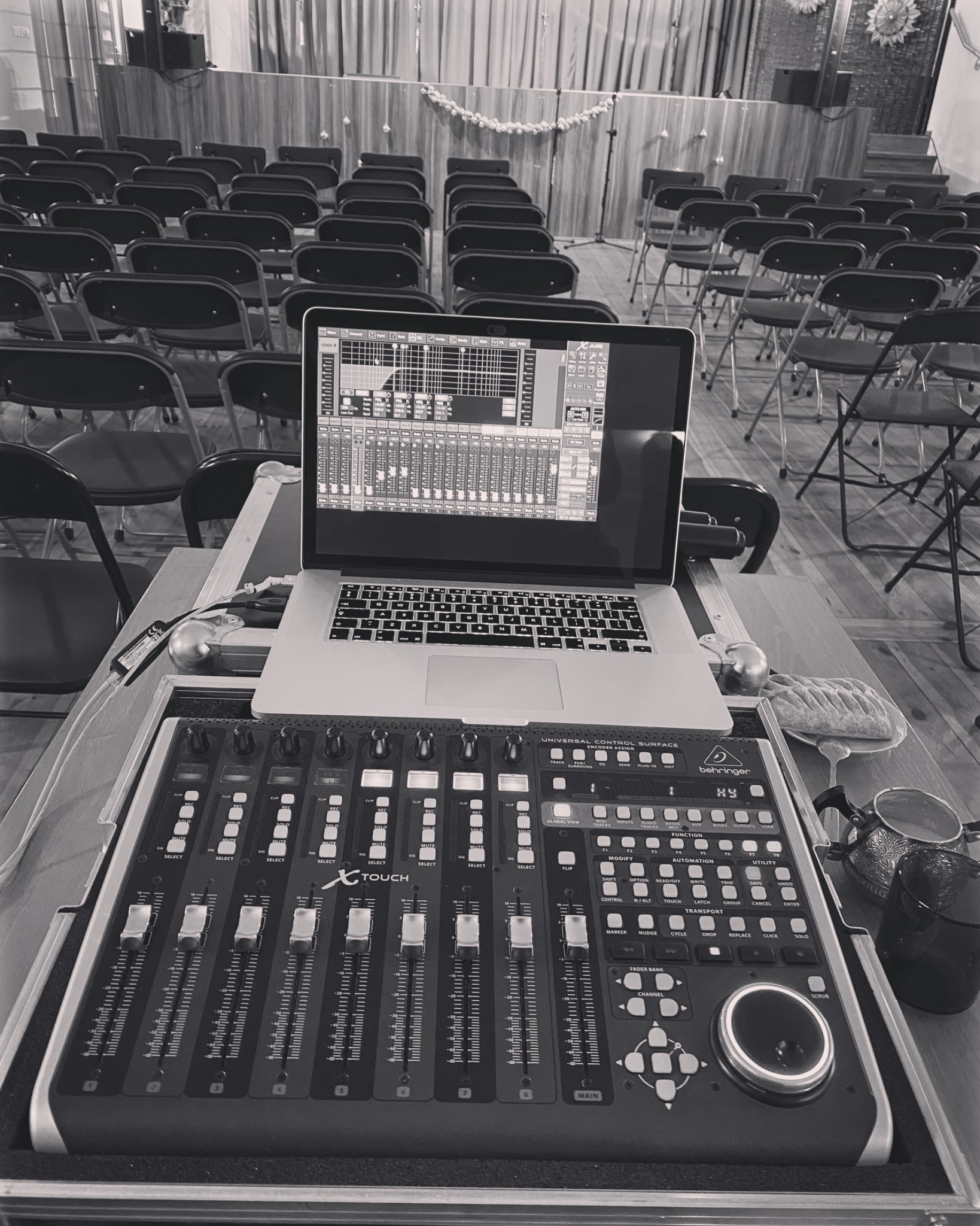 Czarno-białe zdjęcie przedstawia laptop z otwartym programem do miksowania dźwięku, umieszczony na profesjonalnym mikserze Behringer X-Touch, z rzędami krzeseł i dekoracją sceny w tle, sugerujące...