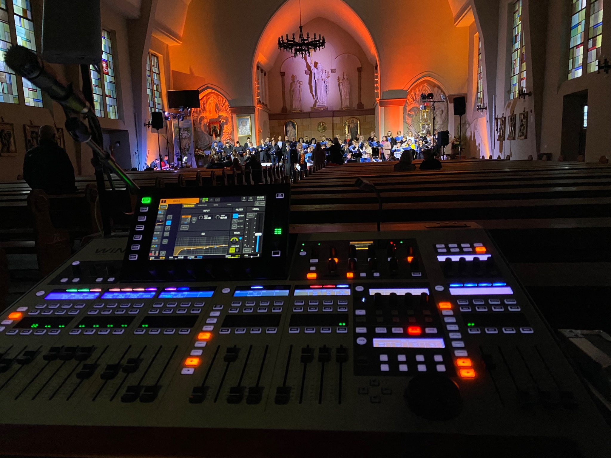 Duży mikser dźwięku z podświetlanymi przyciskami i suwakami, ustawiony w kościele przed chórem i orkiestrą, z widocznymi mikrofonami na statywach.