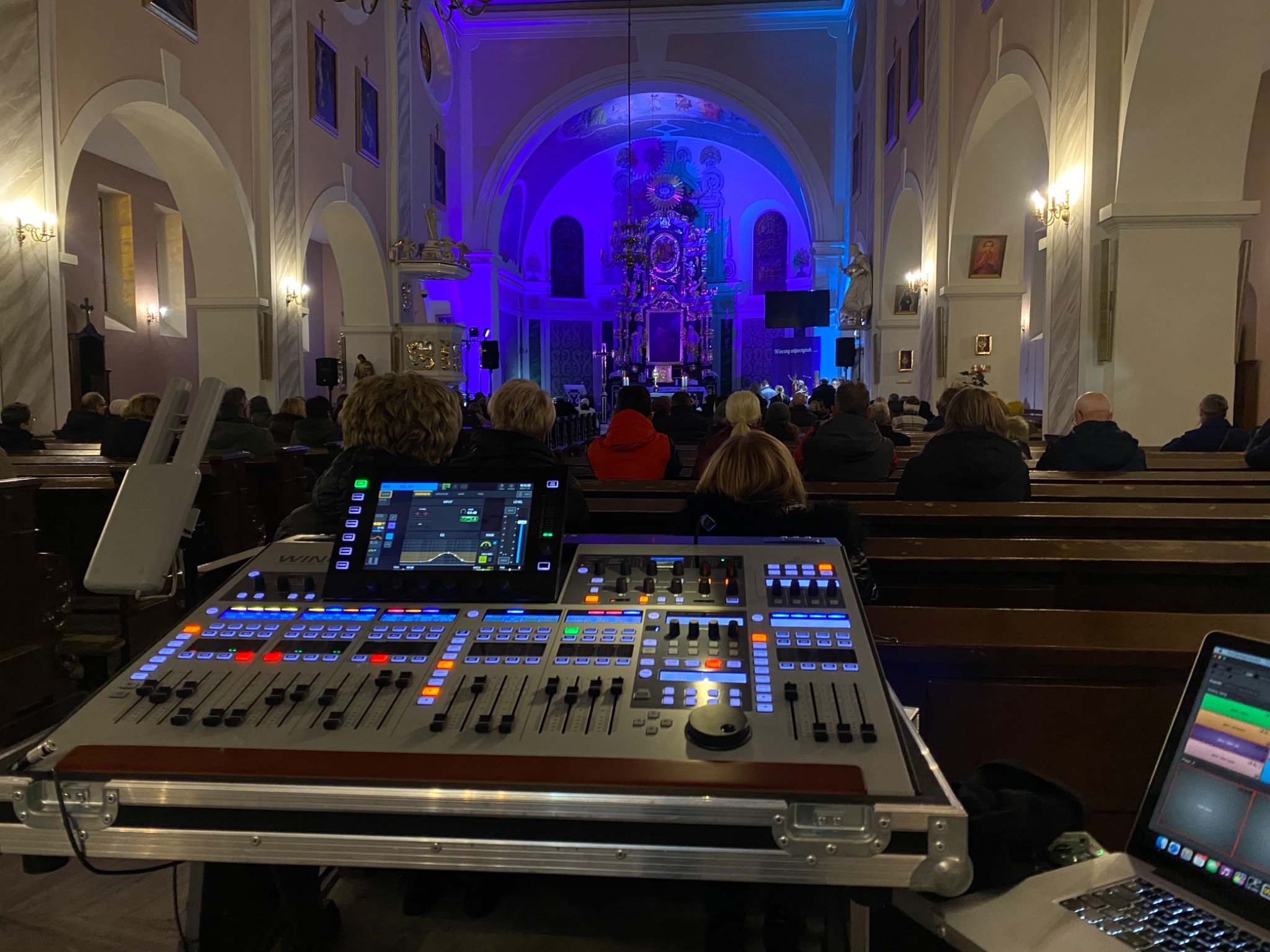 Konsoleta mikserska z laptopem w kościele podczas wydarzenia, widoczna publiczność i niebieskie oświetlenie sceniczne.
