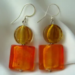 Para ręcznie robionych, pomarańczowo-złotych kolczyków z kwadratowymi i okrągłymi szklanymi koralikami na srebrnych biglach, leżących na białej, satynowej tkaninie.