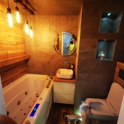 Piotr Falkowski - Nowoczesna łazienka z wanną z hydromasażem, umywalką nablatową, okrągłym lustrem z podświetleniem i podwieszaną toaletą, wykończona płytkami imitującymi drewno i szarym kamieniem.