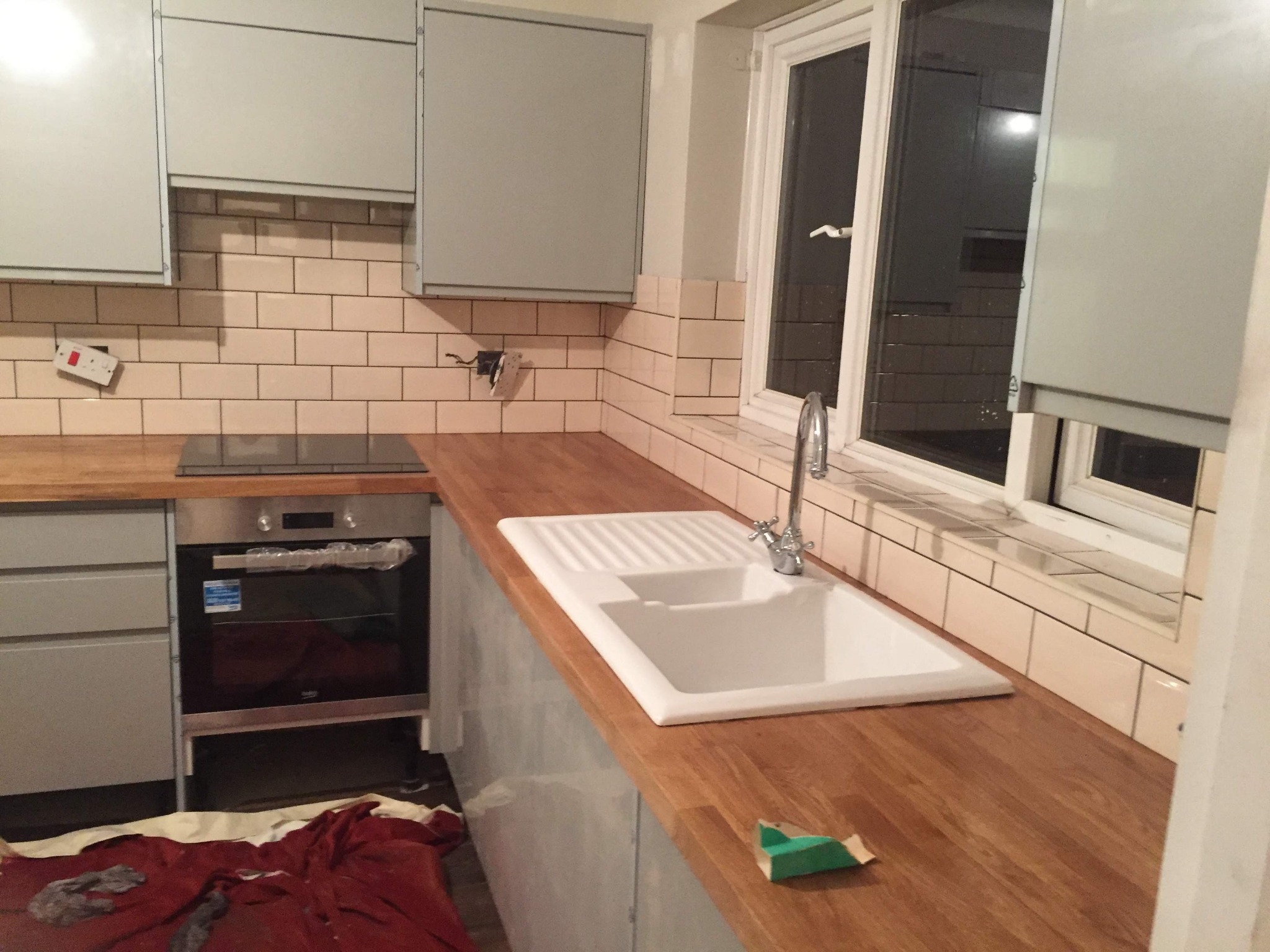 Montaż kuchni: Jasne szafki, drewniany blat z ceramicznym zlewem, beżowe płytki na ścianie, okno z roletą, w tle płyta indukcyjna i piekarnik, na podłodze czerwona folia ochronna.