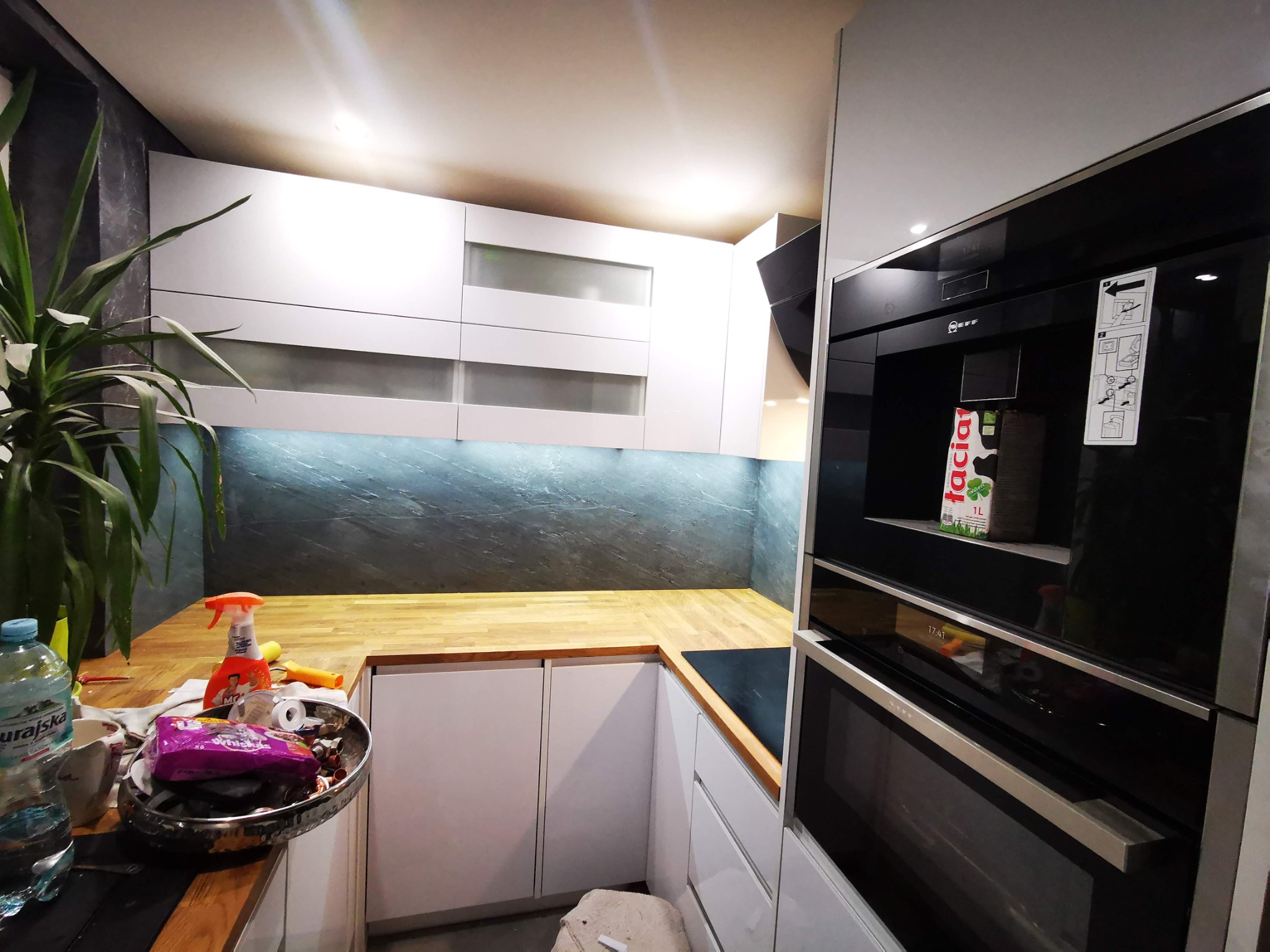 Nowoczesna kuchnia z białymi szafkami, drewnianym blatem i czarnym piekarnikiem Neff. Na blacie widoczny bałagan: butelka wody, środki czystości i miska z drobnymi przedmiotami.