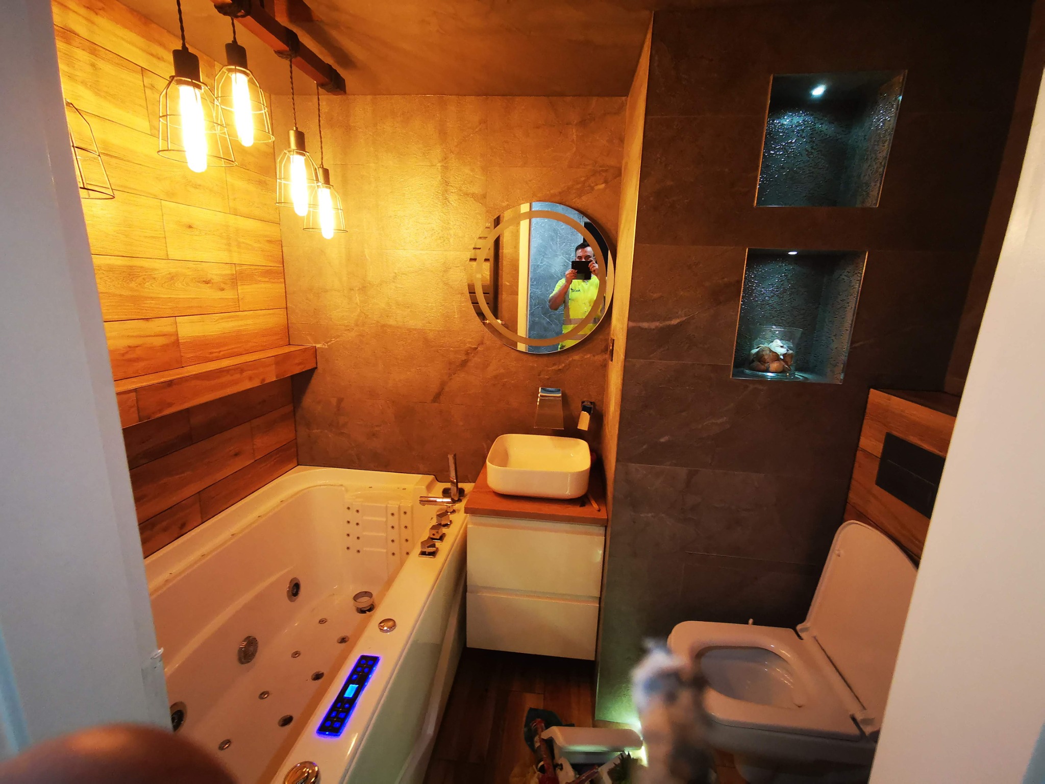 Nowoczesna łazienka z wanną z hydromasażem, umywalką nablatową, okrągłym lustrem z podświetleniem i podwieszaną toaletą, wykończona płytkami imitującymi drewno i szarym kamieniem.