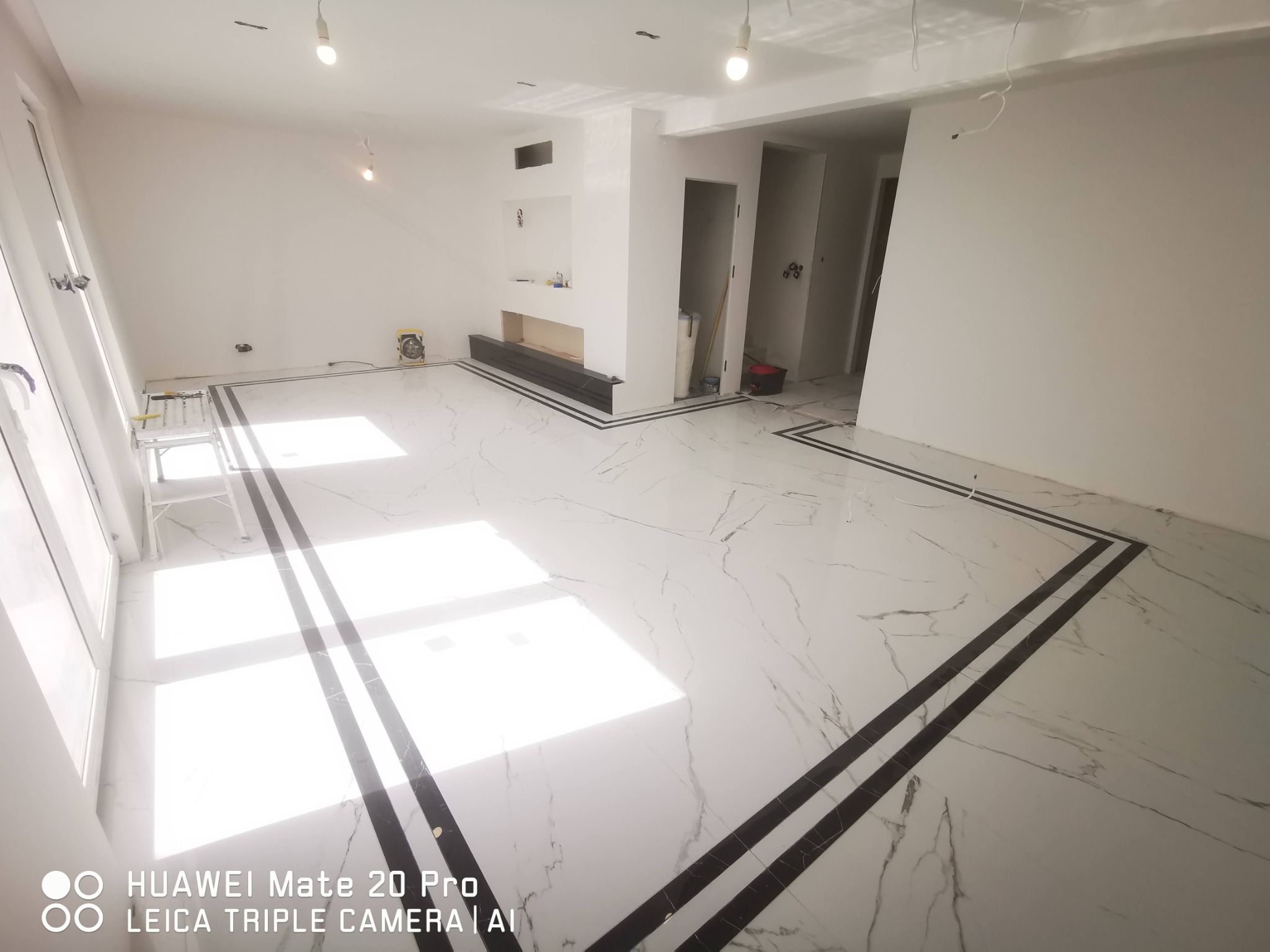 Nowoczesne wnętrze w trakcie prac wykończeniowych: białe ściany, marmurowa podłoga z czarnymi bordiurami, widoczne otwory drzwiowe i przygotowany kominek, drabina malarska w rogu pomieszczenia.