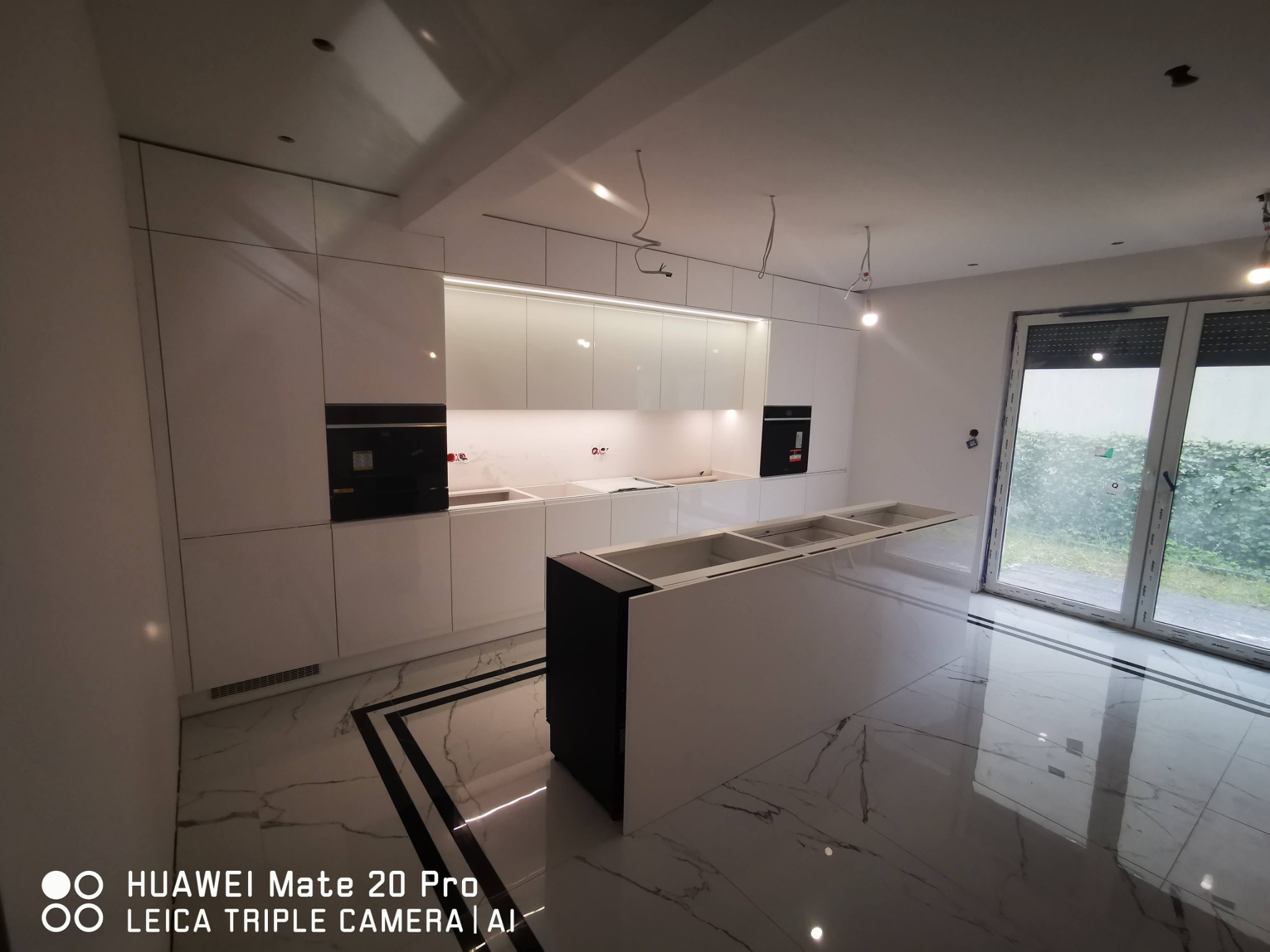 Nowoczesna kuchnia w trakcie wykańczania z białymi, błyszczącymi szafkami, czarnymi piekarnikami i wyspą kuchenną z niezamontowanymi zlewami, marmurowa podłoga z czarnymi akcentami, widoczne...