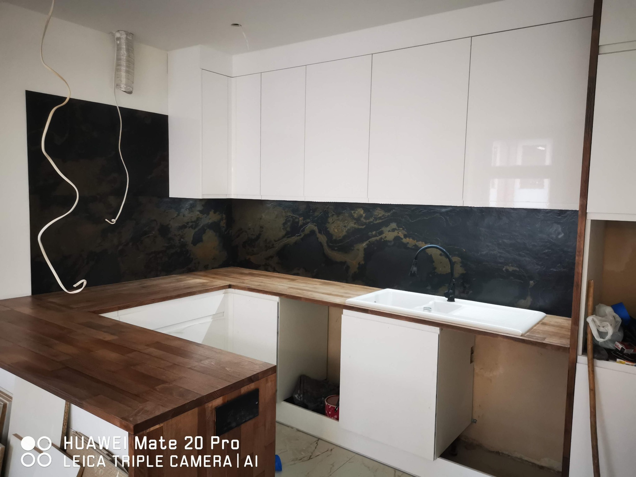 Wnętrze kuchni w trakcie montażu: drewniany blat, białe szafki górne, czarny panel ścienny z marmurkowym wzorem, widoczne niedokończone podłączenia hydrauliczne i elektryczne.