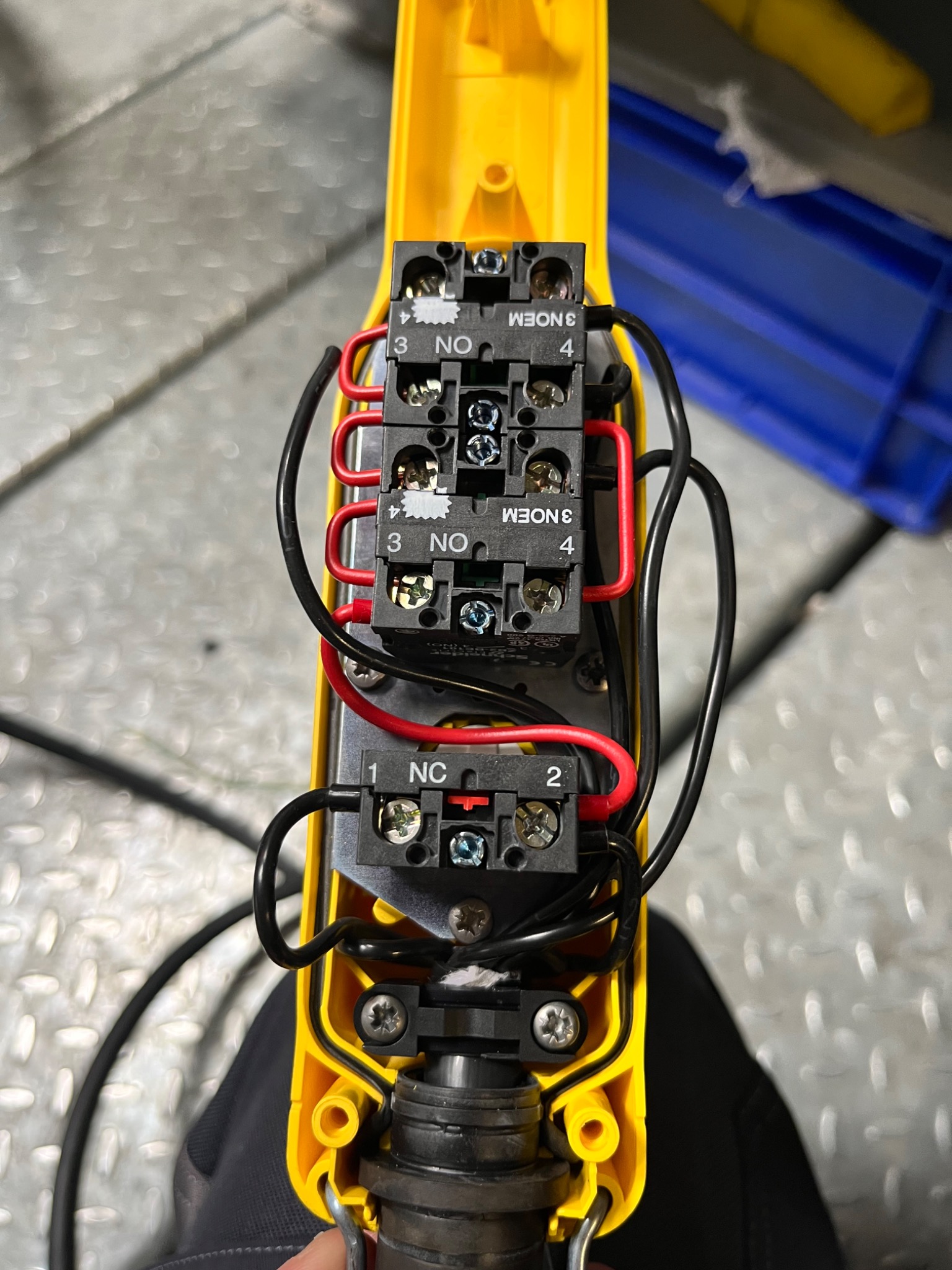 Żółty panel sterowania z podłączonymi przewodami: czarne i czerwone kable wpięte w czarne kostki z oznaczeniami NO i NC, widoczne śruby mocujące.