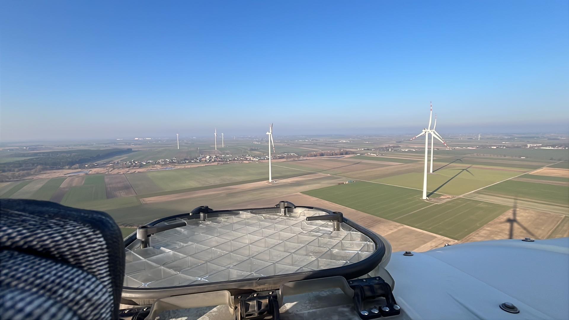 Widok z góry na farmę wiatrową z perspektywy platformy roboczej, oświetlenie LED w kadrze, pola uprawne i błękitne niebo w tle.