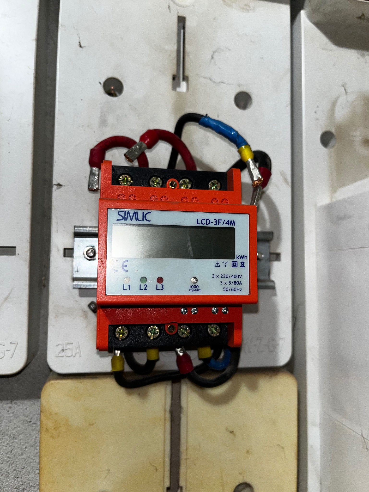 Zamontowany, pomarańczowy licznik energii elektrycznej SIMLIC LCD-3F/4M w białej skrzynce, z podłączonymi przewodami. Widoczne oznaczenia L1, L2, L3 i parametry techniczne.