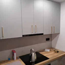 Szare szafki kuchenne z drewnianymi uchwytami, czarny okap Amica, płyta indukcyjna i drewniany blat. Na blacie czajnik, pudełko i narzędzia.