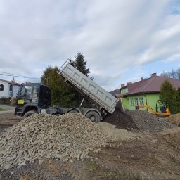 GOSYMAR SPÓŁKA Z OGRANICZONĄ ODPOWIEDZIALNOŚCIĄ - Wyładowywanie kruszywa z wywrotki na placu budowy. W tle domy i mini koparka. Widoczne różne frakcje materiałów. Ujęcie z boku, częściowo na nierównym terenie.