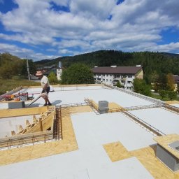 GOSYMAR SPÓŁKA Z OGRANICZONĄ ODPOWIEDZIALNOŚCIĄ - Budowa stropu na białej izolacji zbrojonego wieńca; widoczny pracownik z dmuchawą, drewniane szalunki i elementy zbrojenia na tle gór i błękitnego nieba.