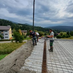 GOSYMAR SPÓŁKA Z OGRANICZONĄ ODPOWIEDZIALNOŚCIĄ - Wylewanie betonu na strop zbrojony w Berezce. Pracownicy budowlani w trakcie pracy z pompą do betonu na tle górskiego krajobrazu i zabudowań.