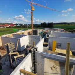 Budowa domu z bloczków betonu komórkowego, widok z góry na fundamenty i ściany parteru, z żurawiem budowlanym w tle i pracownikiem w kasku.