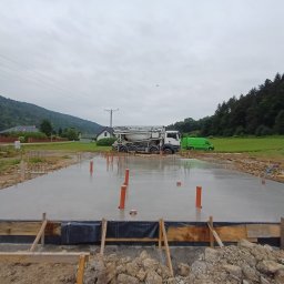 GOSYMAR SPÓŁKA Z OGRANICZONĄ ODPOWIEDZIALNOŚCIĄ - Wylany fundament pod budynek z wystającymi rurami, widoczny betonowóz, w tle góry i zabudowania. Szalunek z desek wzdłuż krawędzi.