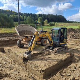 GOSYMAR SPÓŁKA Z OGRANICZONĄ ODPOWIEDZIALNOŚCIĄ - Żółta mini koparka Yanmar i wywrotka na placu budowy, wykop pod fundamenty, widoczne oznaczenia wykopu, w tle drzewa i niebo z chmurami.