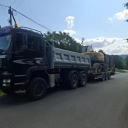 Ciężarówka MAN z naczepą przewożąca koparkę kołową na tle błękitnego nieba z chmurami. Widoczne drzewa i słupy energetyczne.