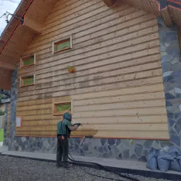 Pracownik w kombinezonie ochronnym piaskuje drewnianą elewację domu. Widoczne zabezpieczone okna i kamienny cokół. Częściowo oczyszczona ściana z desek.