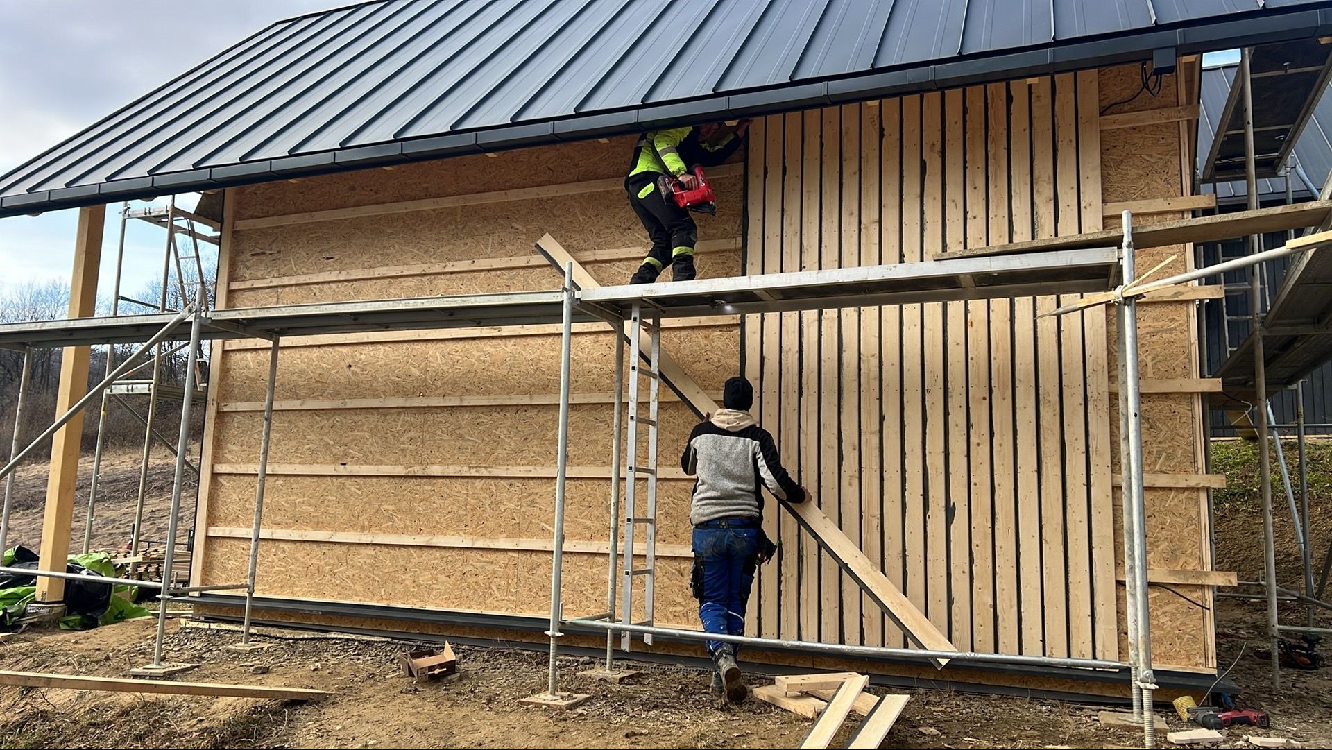 Montaż elewacji drewnianej na budynku w trakcie budowy. Dwóch pracowników na rusztowaniu, jeden mocuje deski, drugi podaje. Widoczne płyty OSB i czarny dach.