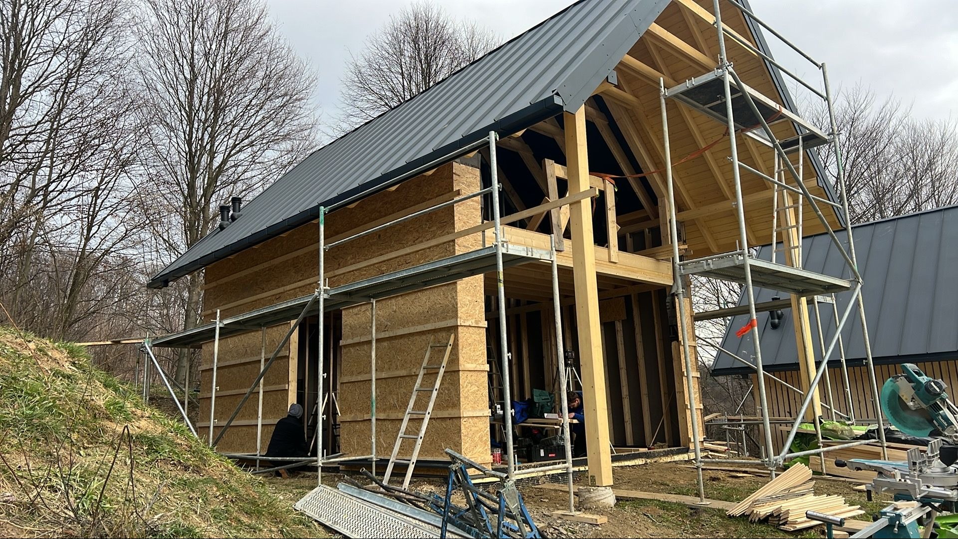 Budowa domu szkieletowego: widok z zewnątrz na konstrukcję w trakcie montażu, z rusztowaniem, elementami drewnianymi i częściowo ukończonym dachem.