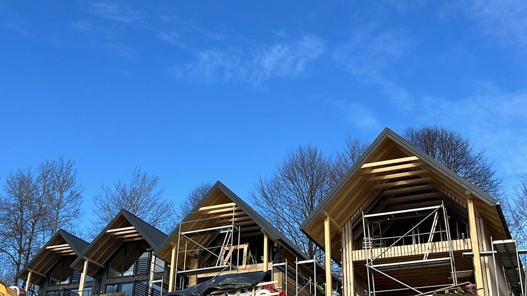 Trzy domy w stanie surowym otwartym, z drewnianą konstrukcją dachu i rusztowaniami, na tle błękitnego nieba. Jeden dom ma już zamontowane okna i elewację.