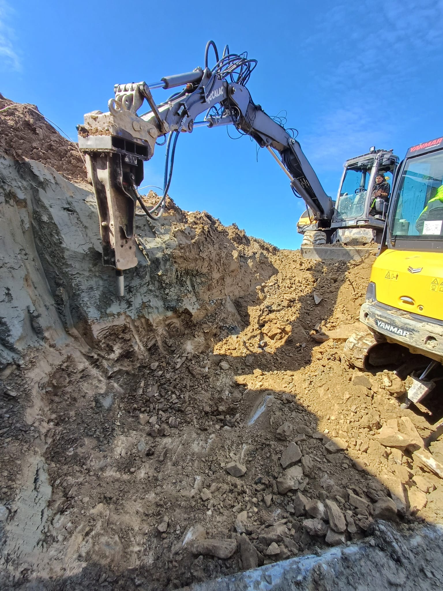 Żółta koparka Yanmar z operatorem w kabinie, rozbija skałę hydraulicznym młotem Mecalac na tle błękitnego nieba, tworząc wykop pod instalacje.