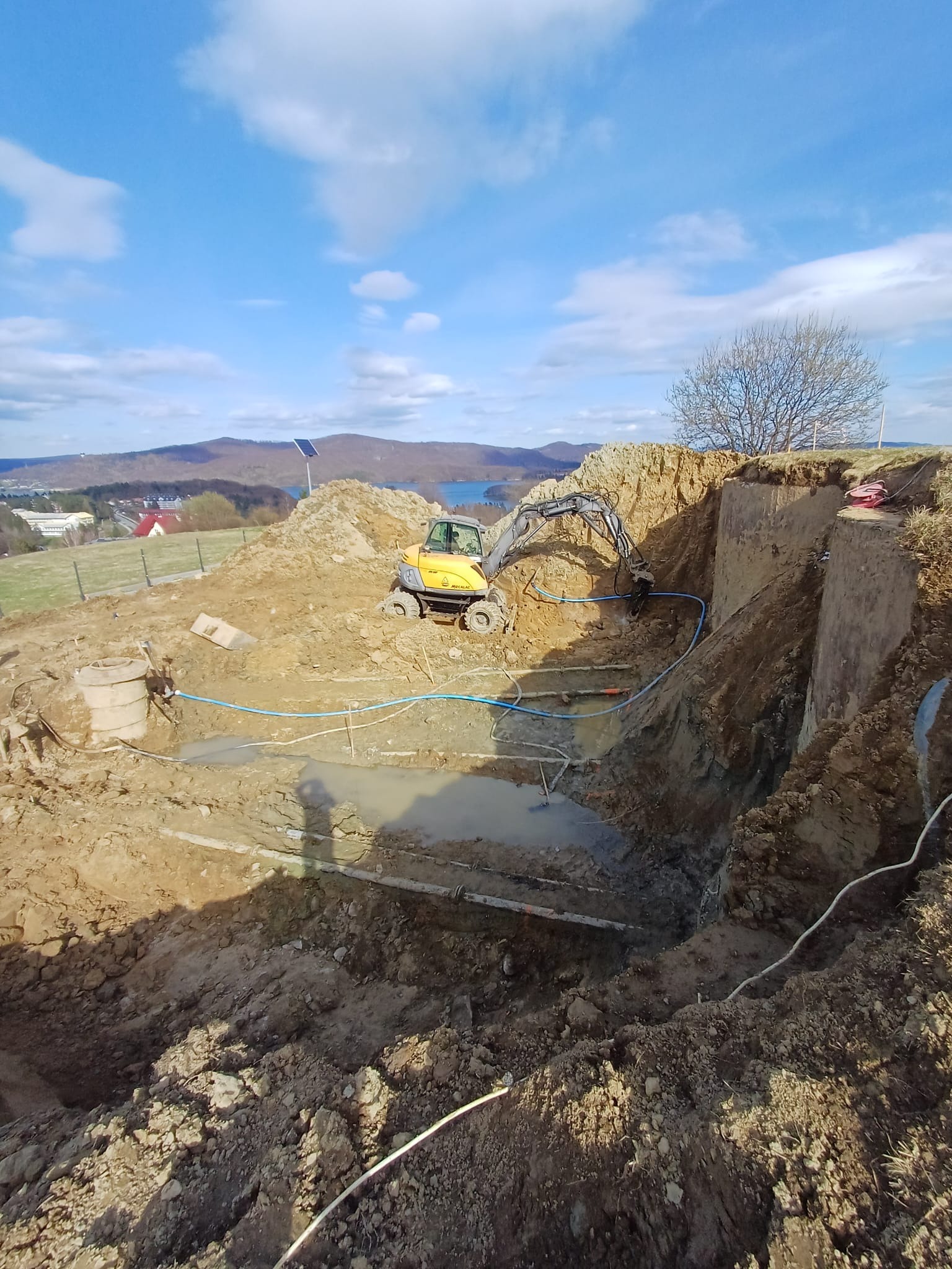 Żółta koparka pracuje w wykopie ziemnym z widocznymi rurami, na tle błękitnego nieba z chmurami i odległego krajobrazu z jeziorem i górami.