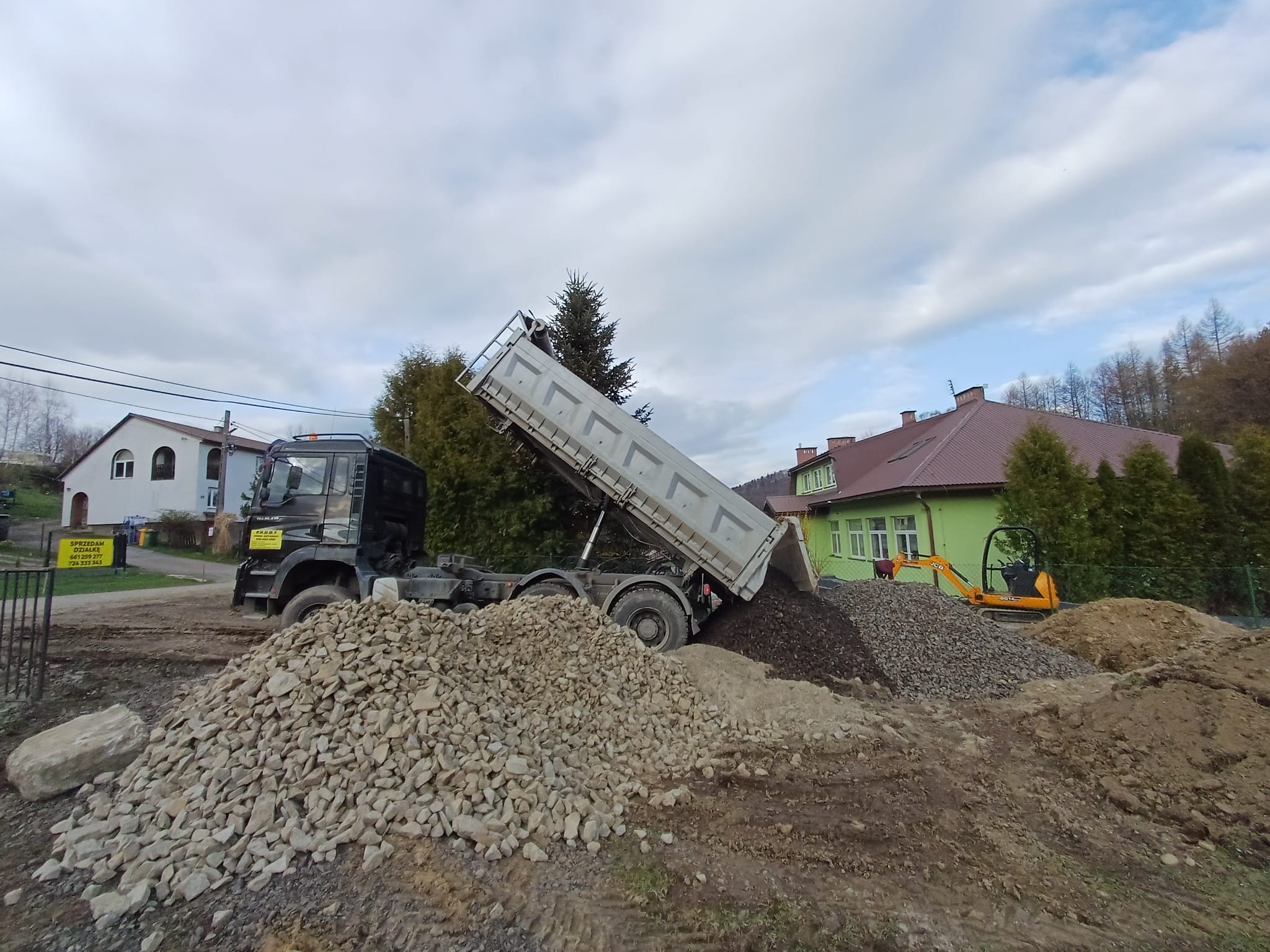 Wyładowywanie kruszywa z wywrotki na placu budowy. W tle domy i mini koparka. Widoczne różne frakcje materiałów. Ujęcie z boku, częściowo na nierównym terenie.