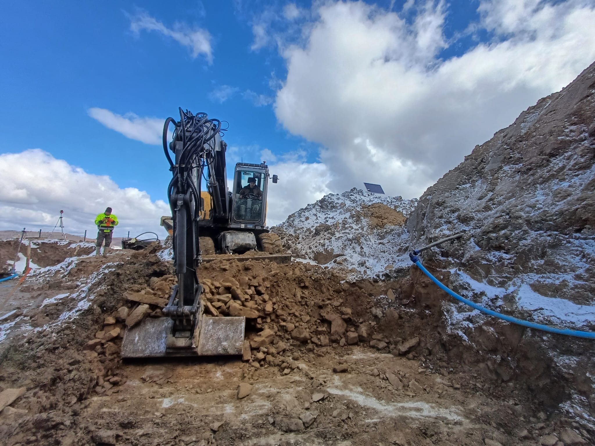Żółta koparka w trakcie prac ziemnych na placu budowy, widoczny pracownik w odblaskowej kamizelce i niebieska rura. W tle błękitne niebo z chmurami.