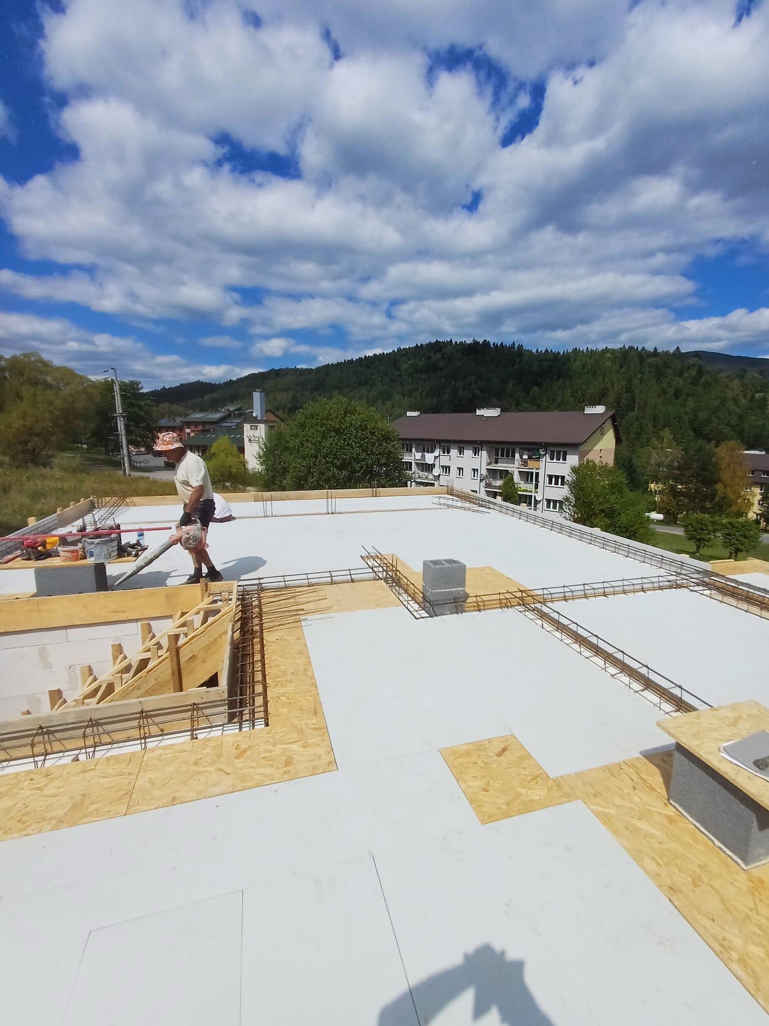 Budowa stropu na białej izolacji zbrojonego wieńca; widoczny pracownik z dmuchawą, drewniane szalunki i elementy zbrojenia na tle gór i błękitnego nieba.