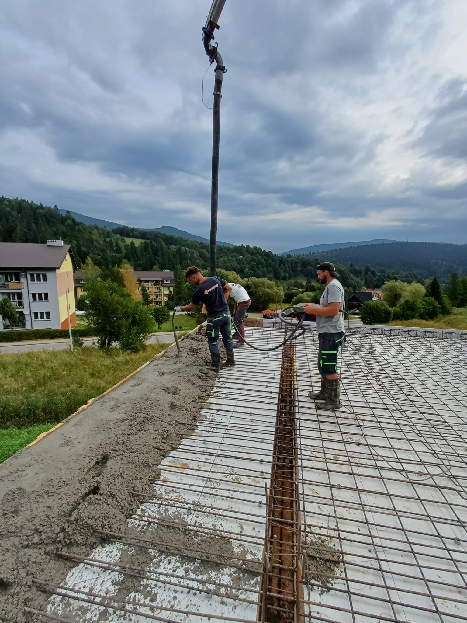 Wylewanie betonu na strop zbrojony w Berezce. Pracownicy budowlani w trakcie pracy z pompą do betonu na tle górskiego krajobrazu i zabudowań.