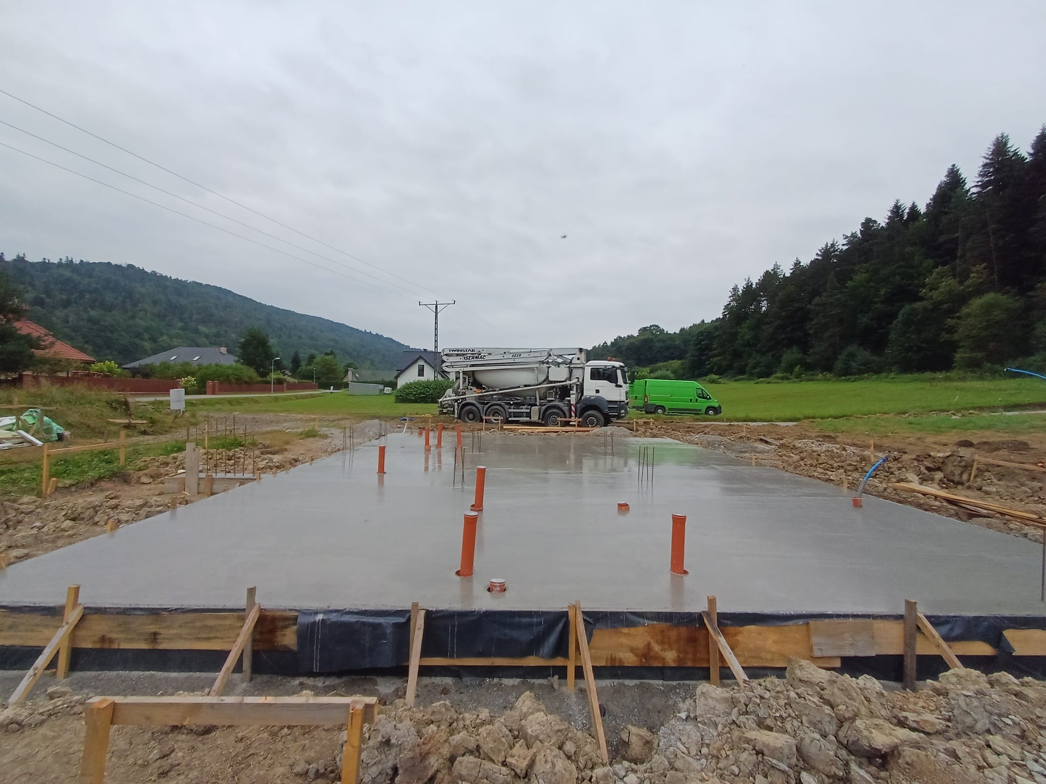 Wylany fundament pod budynek z wystającymi rurami, widoczny betonowóz, w tle góry i zabudowania. Szalunek z desek wzdłuż krawędzi.
