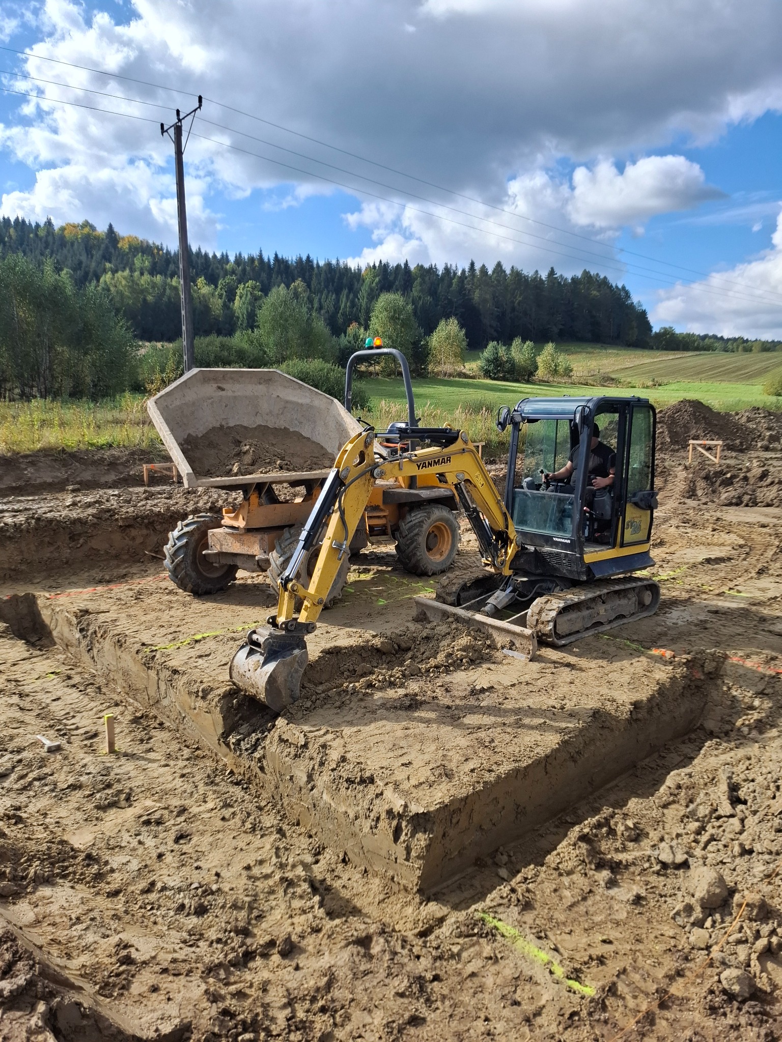 Żółta mini koparka Yanmar i wywrotka na placu budowy, wykop pod fundamenty, widoczne oznaczenia wykopu, w tle drzewa i niebo z chmurami.
