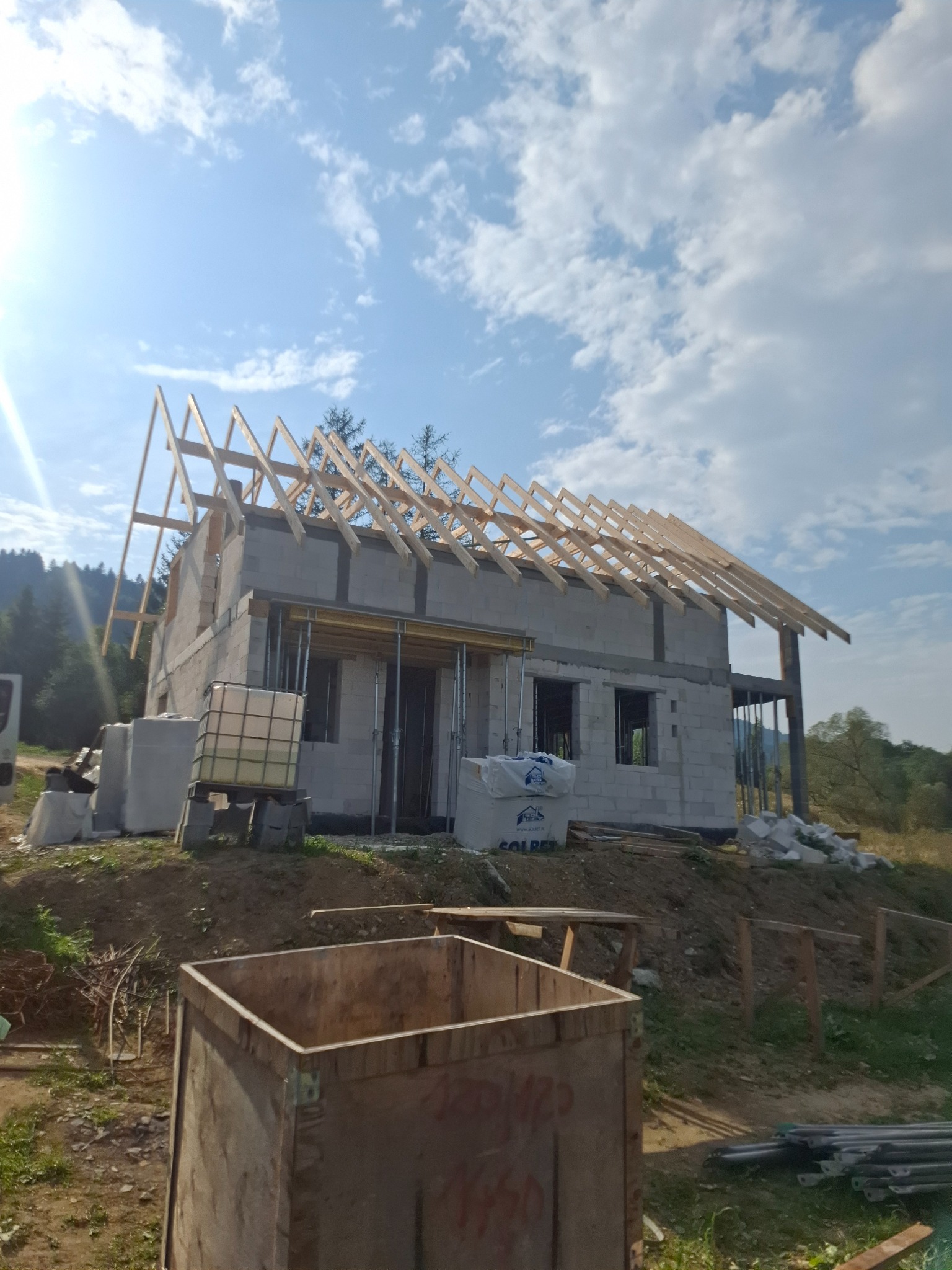 Wznoszenie domu z bloczków: surowa konstrukcja z drewnianą więźbą dachową na tle błękitnego nieba z chmurami. Widoczne materiały budowlane i elementy szalunków.