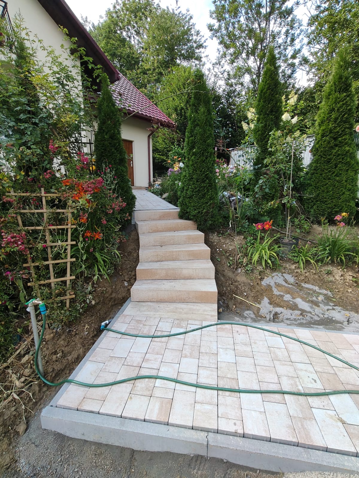 Ułożona kostka brukowa tworząca ścieżkę i schody prowadzące do domu. Widoczne rośliny, kwiaty i trawniki. W tle budynek z czerwonym dachem.