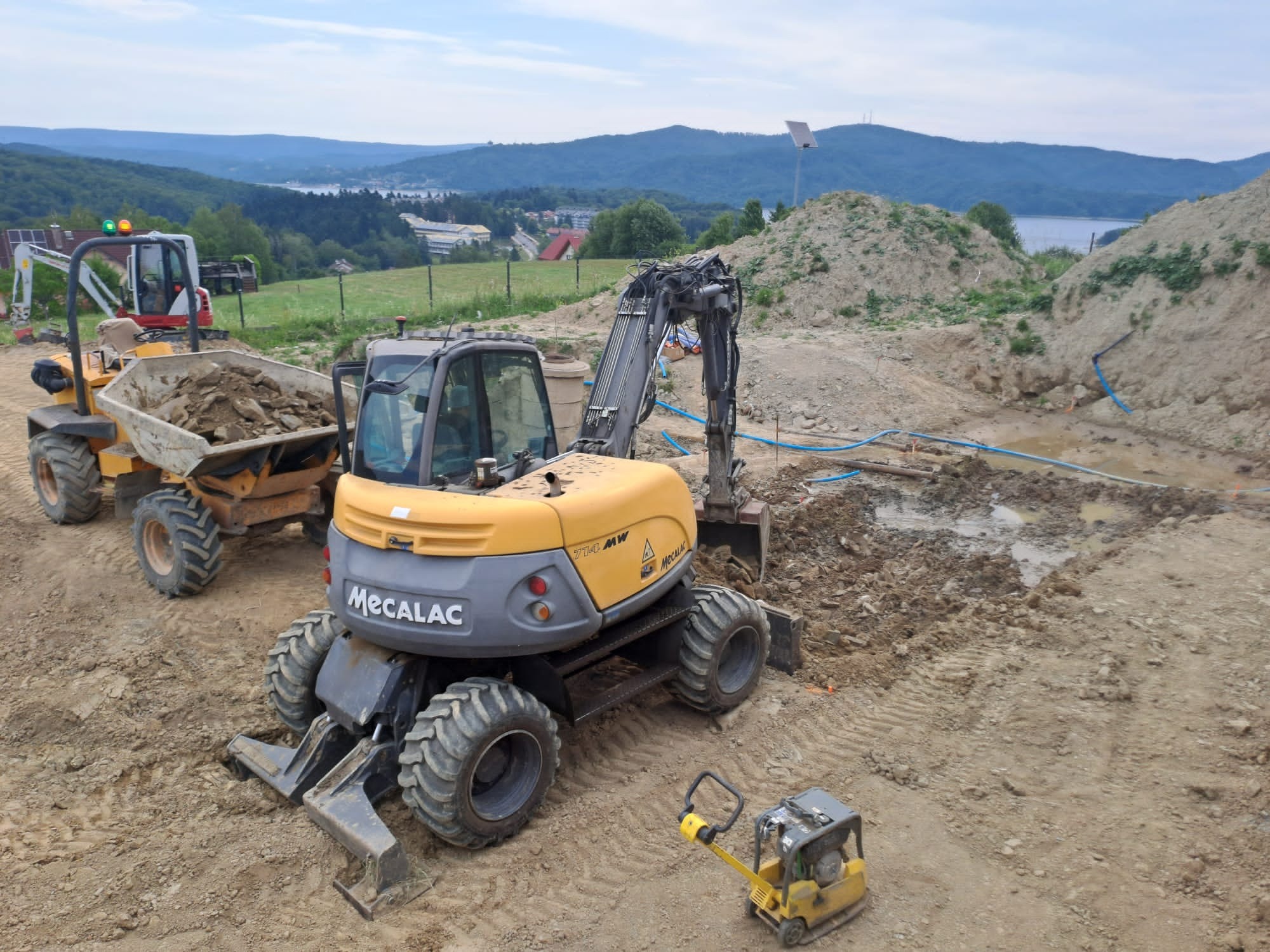 Żółto-szara koparka Mecalac 714MW na placu budowy, z widocznymi śladami pracy w ziemi i w oddali wozidło wypełnione gruzem, krajobraz górski w tle.