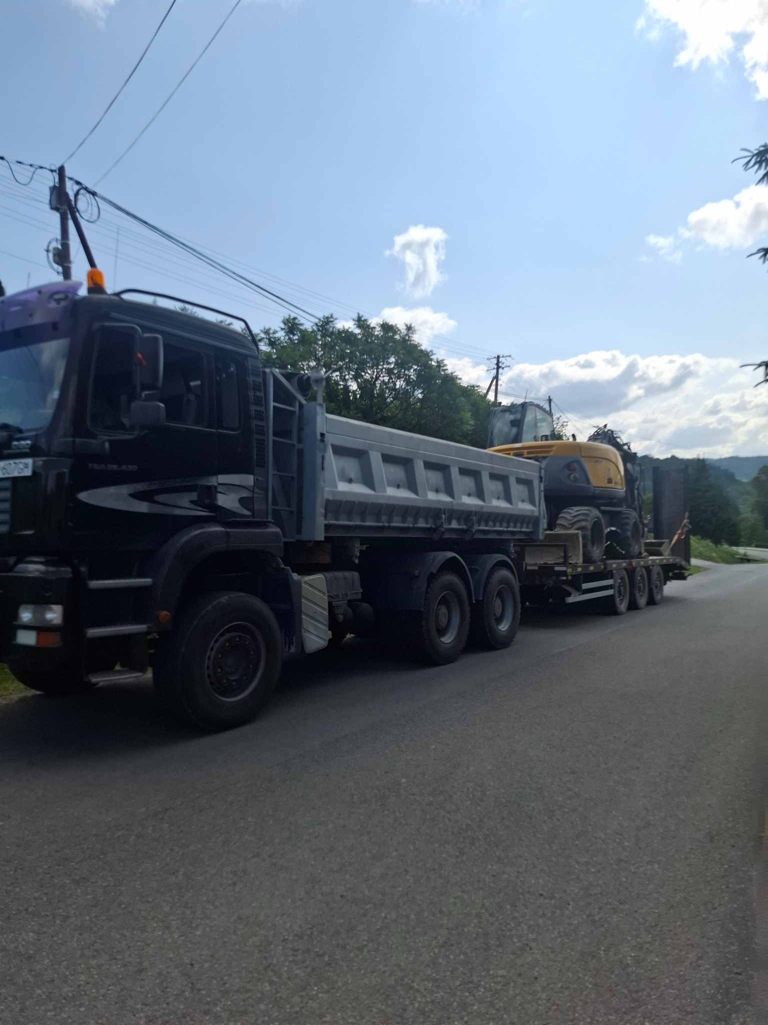 Ciężarówka MAN z naczepą przewożąca koparkę kołową na tle błękitnego nieba z chmurami. Widoczne drzewa i słupy energetyczne.