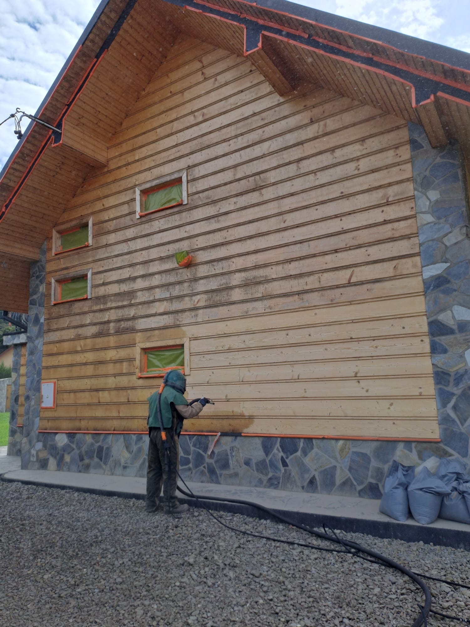 Pracownik w kombinezonie ochronnym piaskuje drewnianą elewację domu. Widoczne zabezpieczone okna i kamienny cokół. Częściowo oczyszczona ściana z desek.