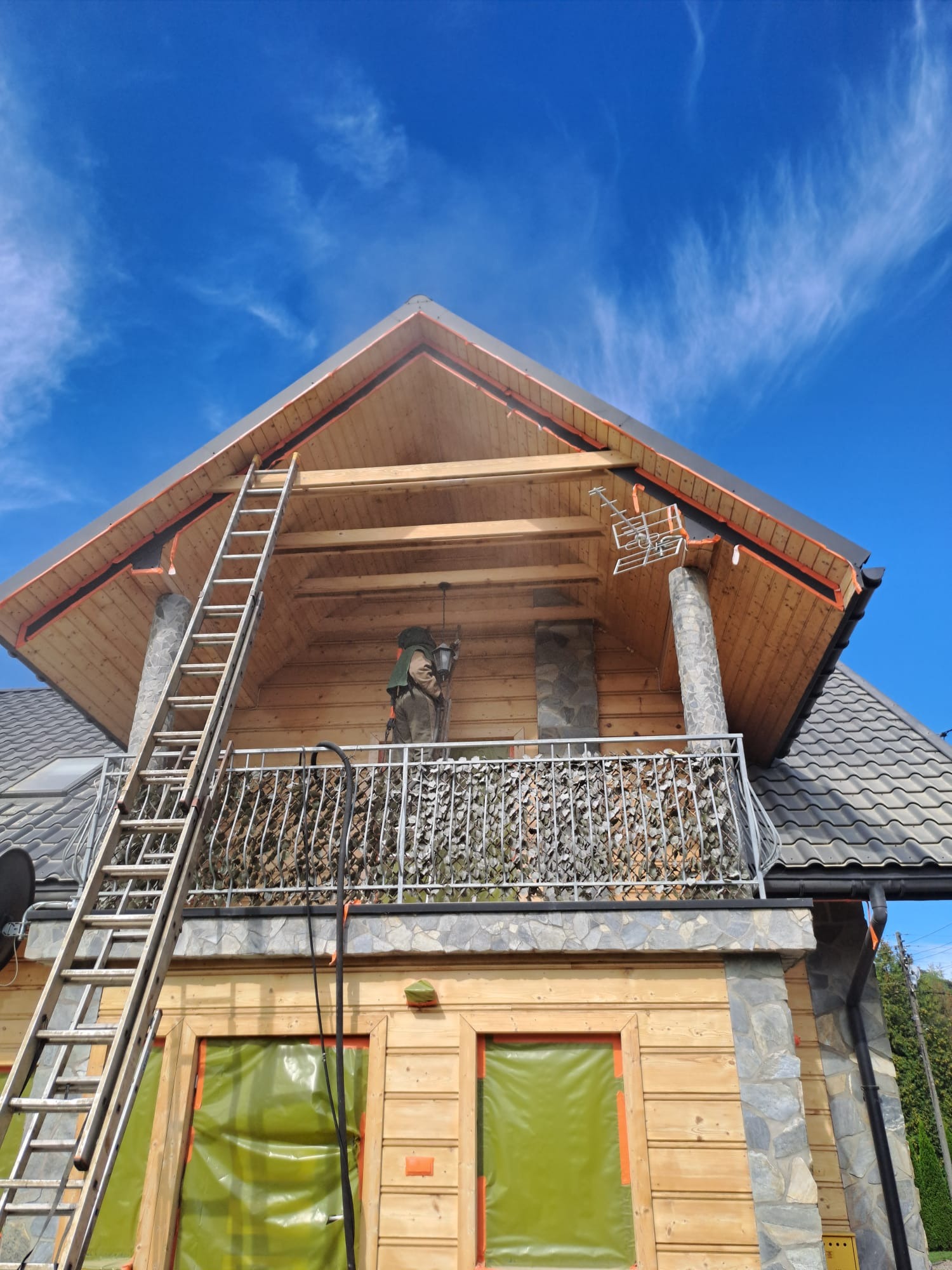 Drewniany dom z balkonem i kamiennymi elementami w trakcie budowy. Widoczna drabina, zabezpieczone okna folią i pracownik na balkonie. Błękitne niebo w tle.