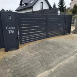 Nowoczesna brama wjazdowa z szarego metalu, z numerem domu 'Gacka 126' i domofonem, na tle kostki brukowej i domu z ciemnym dachem.