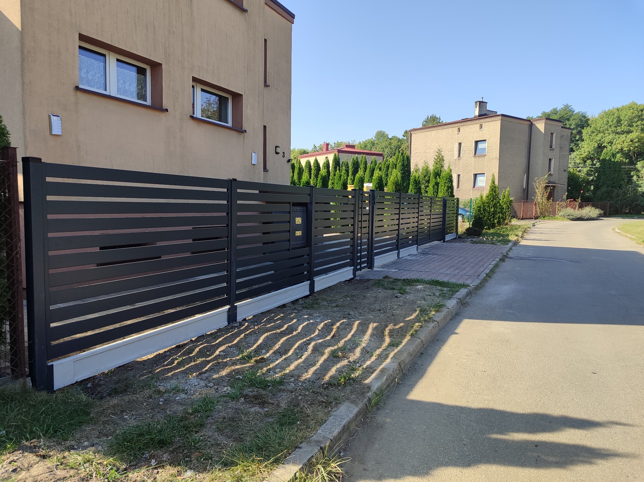 Nowoczesne, poziome ogrodzenie metalowe w kolorze antracytowym, z betonową podmurówką, wzdłuż chodnika i budynków mieszkalnych.