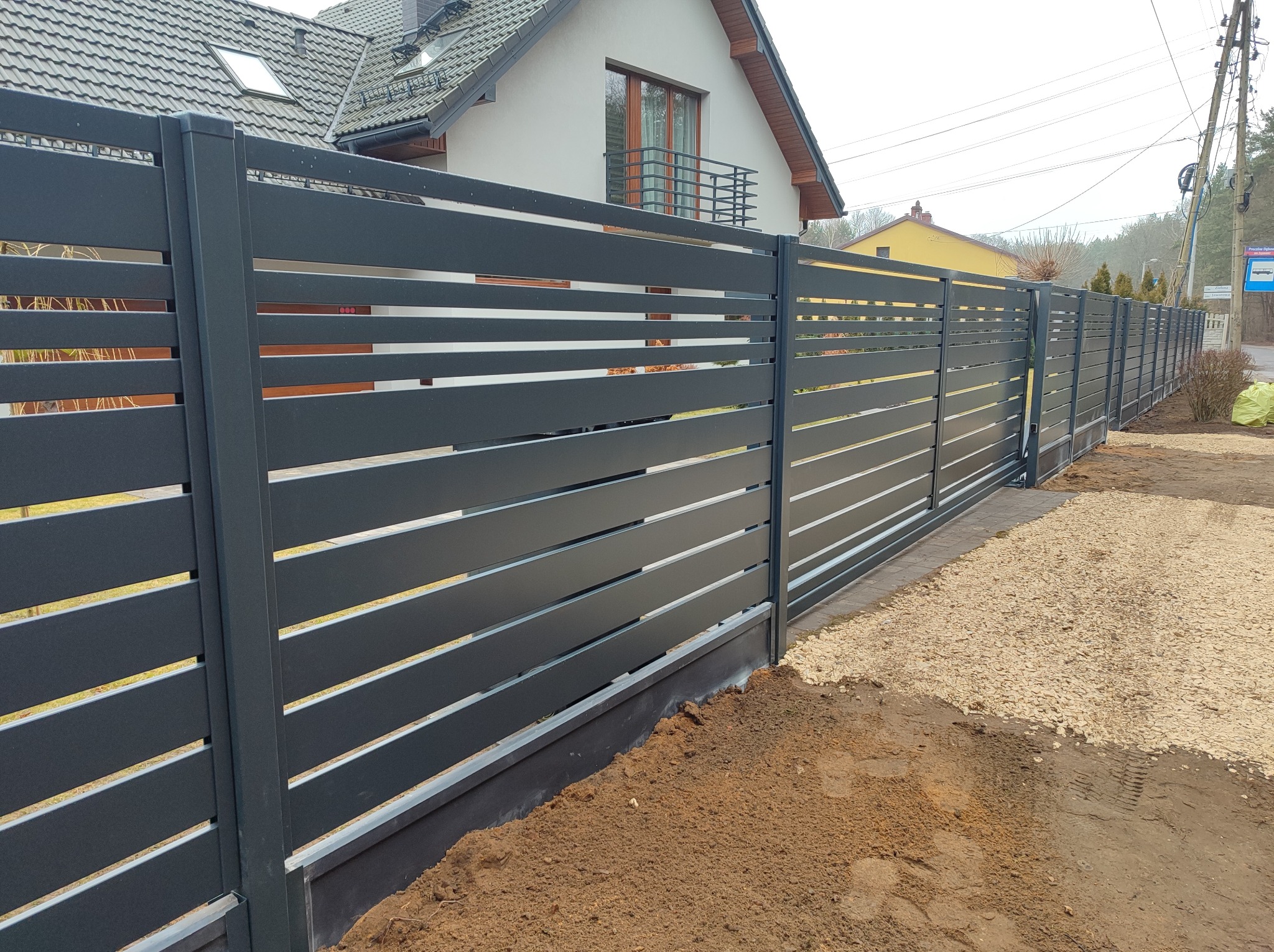 Nowoczesne, antracytowe ogrodzenie metalowe z poziomymi panelami, widok z zewnątrz, świeżo po montażu, z widoczną ziemią i kamieniami wokół.