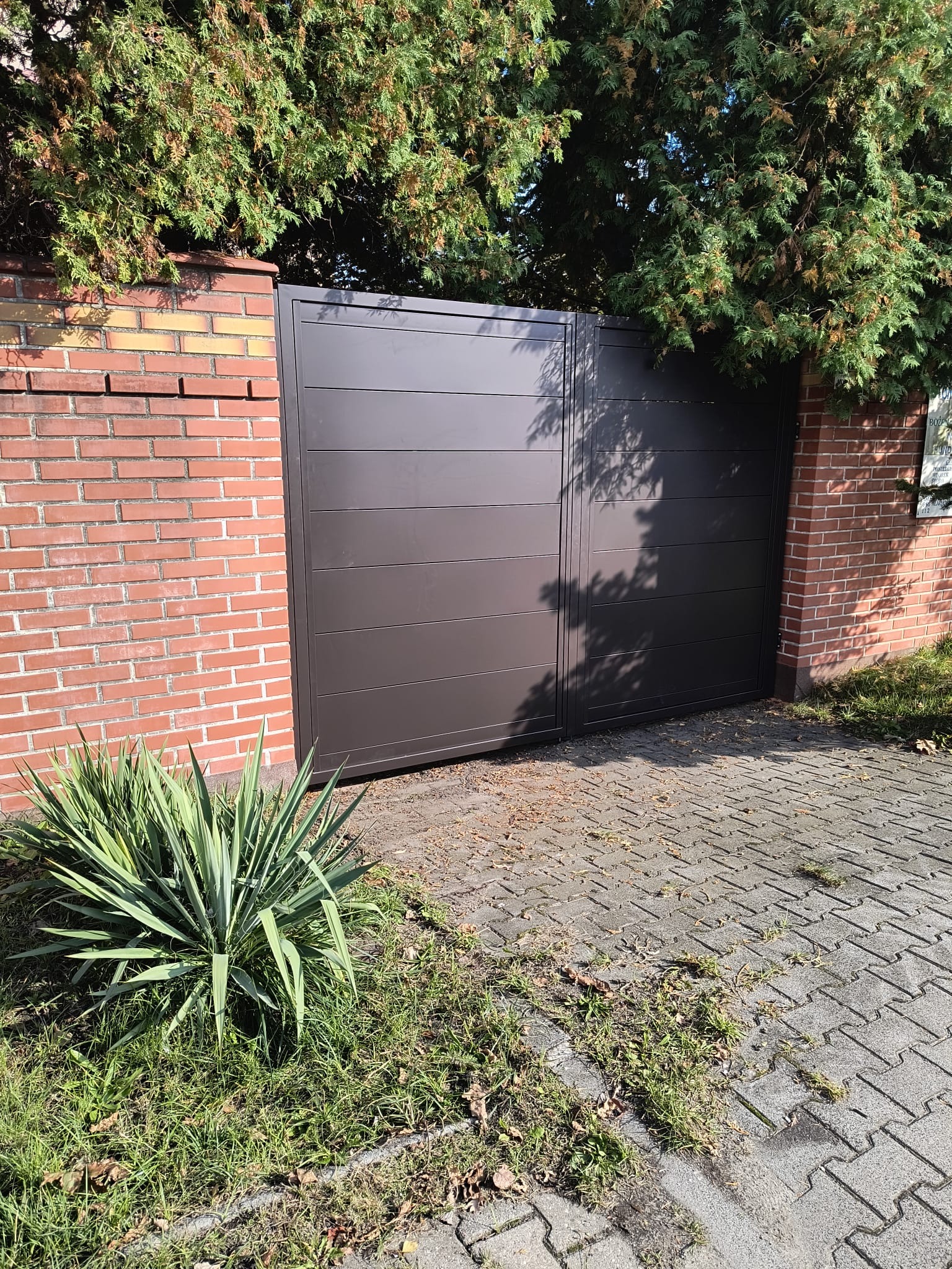 Nowoczesna, brązowa brama wjazdowa z poziomymi panelami, osadzona między ceglanymi słupkami, z zielenią w tle i kostką brukową na podjeździe.