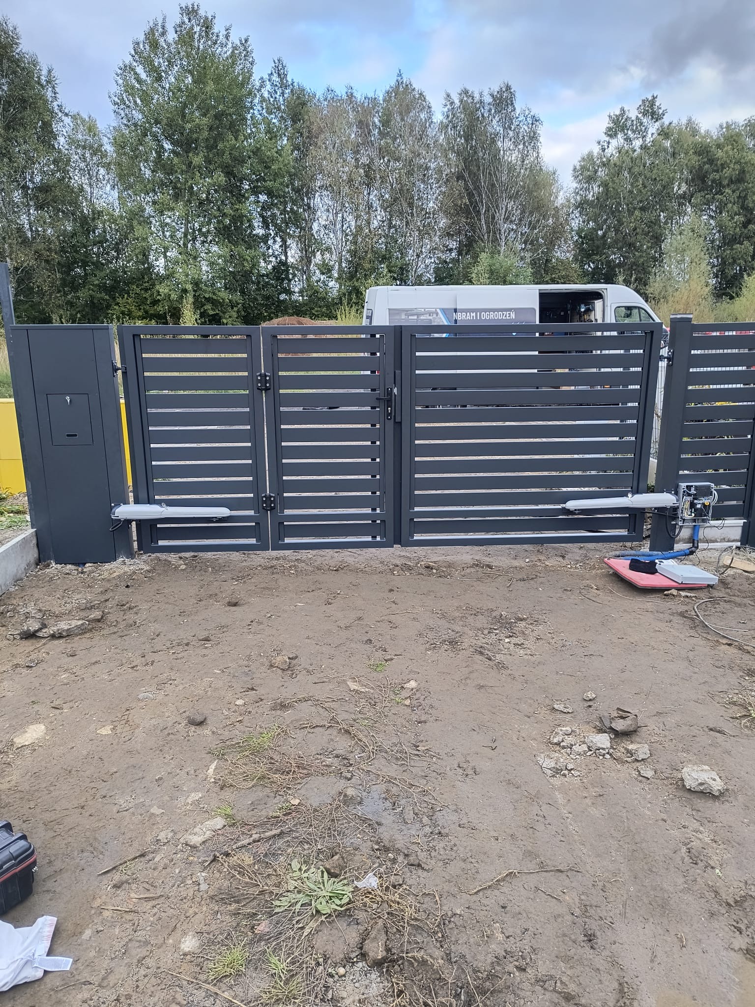 Nowoczesna, grafitowa brama wjazdowa z automatyką na posesji. Widoczny częściowo bus firmy montującej bramy. Teren wokół bramy częściowo przygotowany.