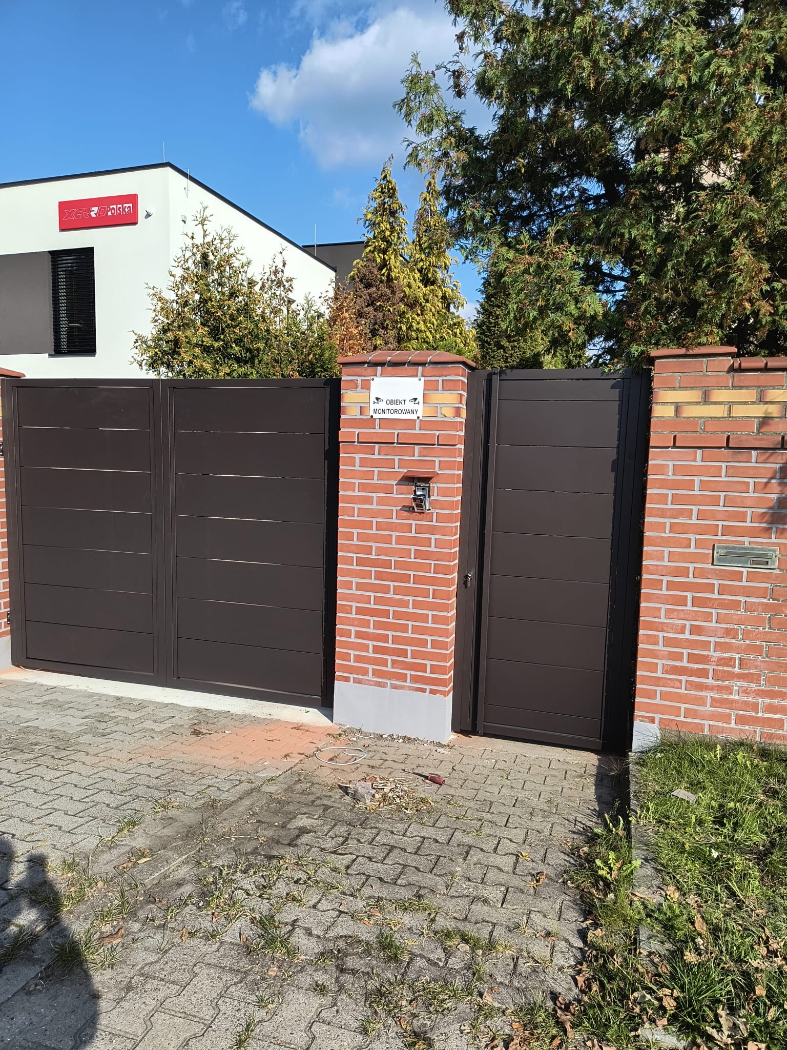 Nowoczesna, brązowa brama wjazdowa z furtką, zamontowana w ceglanym ogrodzeniu. Widoczny budynek z logo firmy w tle i kostka brukowa na podjeździe.